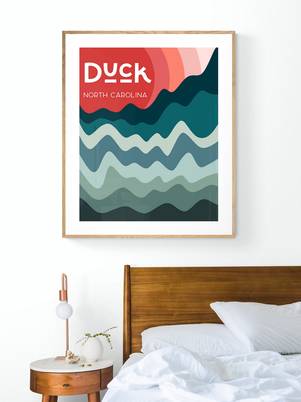 Modern art print featuring layered wavy bands in teal, blue, and coral hues with text 'Duck North Carolina,' displayed above a wooden bed and round bedside table.