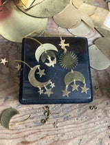 Four brass celestial ornaments including crescent moons with stars, a sunburst, and a bird on a dark square dish surrounded by other brass shapes on wood.