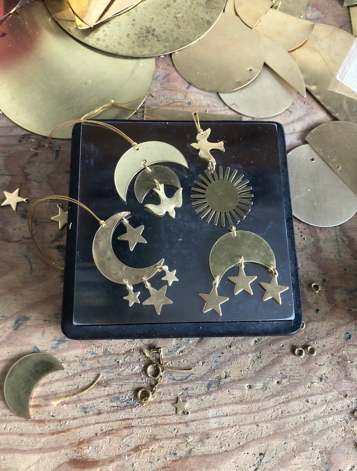 Four brass celestial ornaments including crescent moons with stars, a sunburst, and a bird on a dark square dish surrounded by other brass shapes on wood.