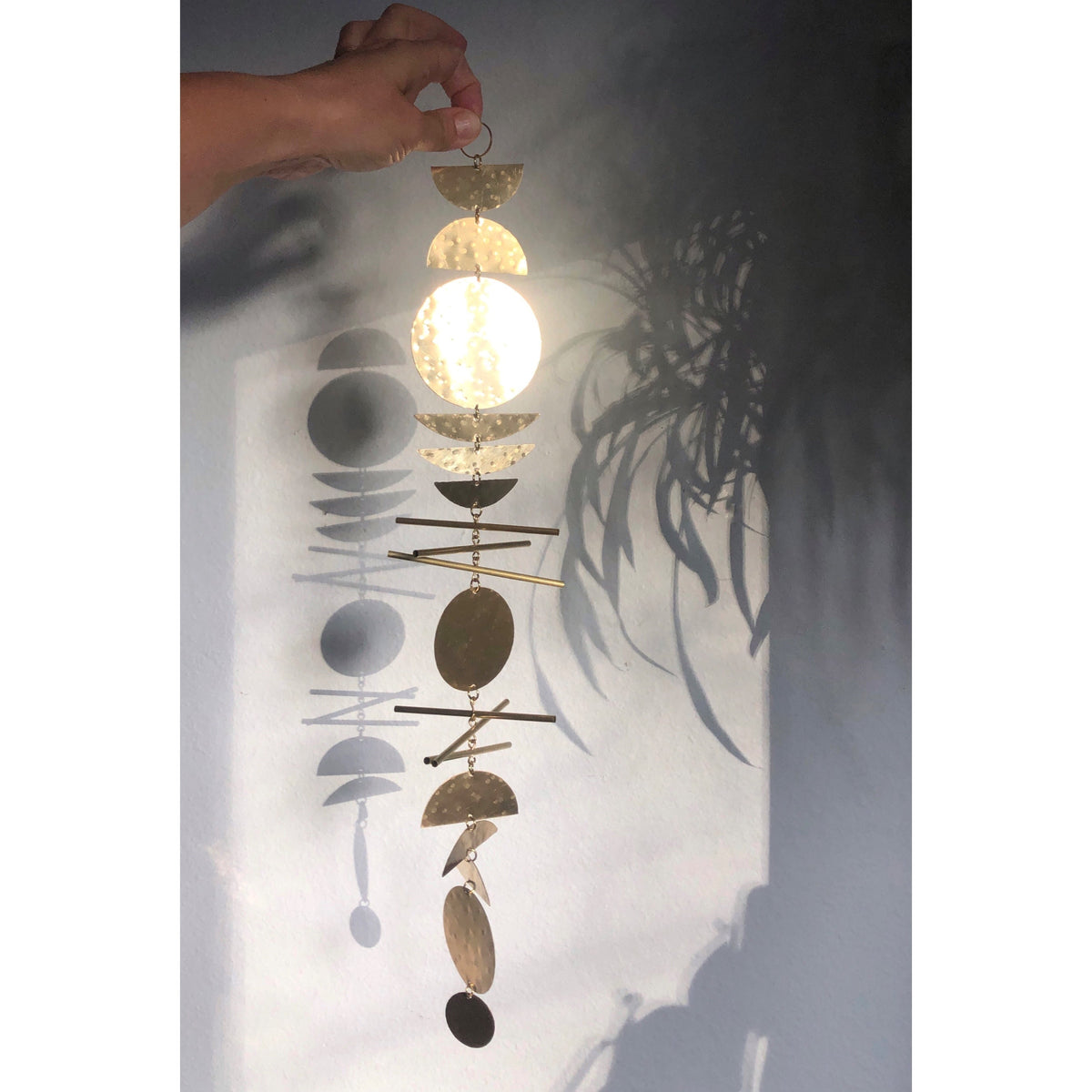 Hand holding a gold-toned metal wall hanging with geometric half circles, circles, and rods casting shadows on a gray wall with a plant shadow.