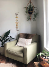 Beige armchair with a furry brown cushion and light beige pillow, placed between two green plants in a light-filled corner with a hanging wooden mobile on the wall.
