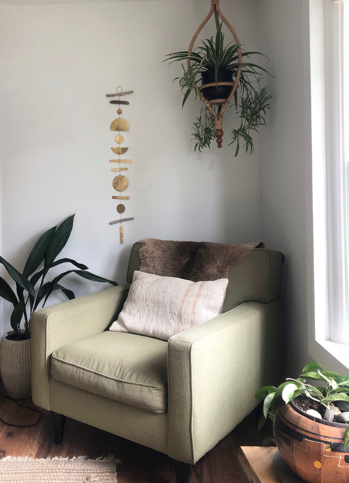 Beige armchair with a furry brown cushion and light beige pillow, placed between two green plants in a light-filled corner with a hanging wooden mobile on the wall.