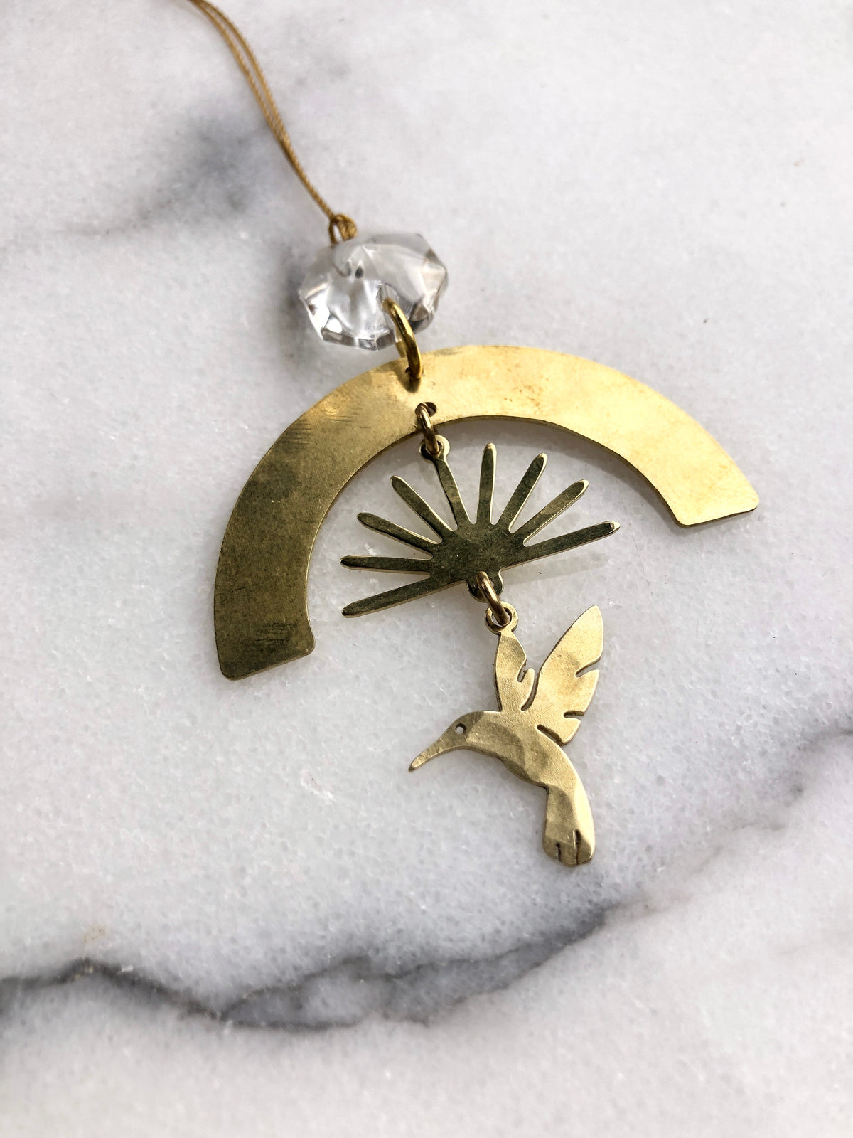 Golden metal ornament featuring a sunburst shape suspended under a crescent arc with a small hummingbird charm hanging below, connected to a clear faceted crystal bead.