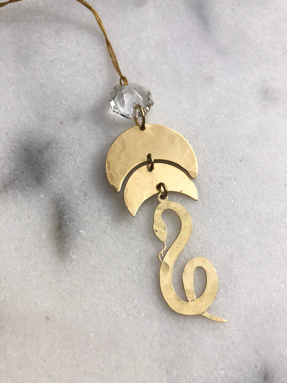 Hammered gold-tone metal serentine shaped ornament with linked crescent shapes and a curled tail, topped with a faceted clear vintage glass crystal bead on a gold loop.