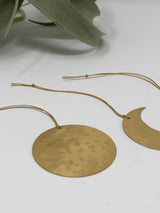 Two hammered brass celestial ornaments, one round and one crescent-shaped, suspended by thin beige cords, displayed on a white surface with a leafy plant in the background.