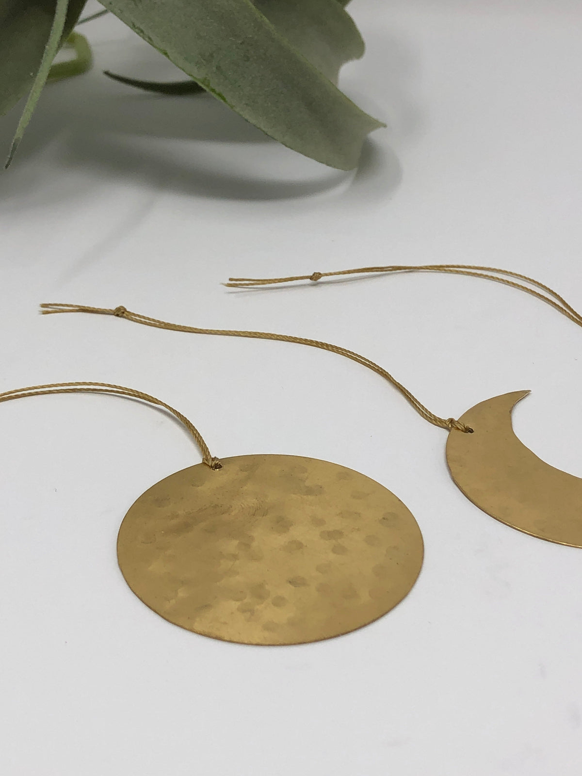 Two hammered brass celestial ornaments, one round and one crescent-shaped, suspended by thin beige cords, displayed on a white surface with a leafy plant in the background.
