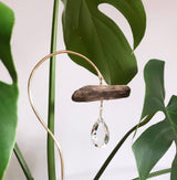 Gold curved metal stand holding a piece of driftwood with a teardrop crystal hanging beneath, surrounded by large green Monstera leaves against a white background