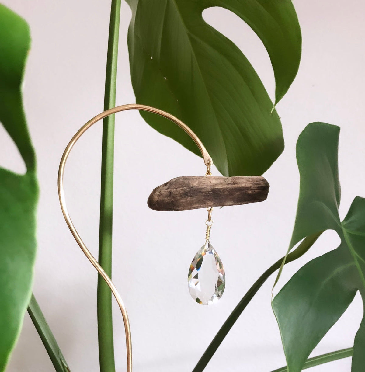 Gold curved metal stand holding a piece of driftwood with a teardrop crystal hanging beneath, surrounded by large green Monstera leaves against a white background