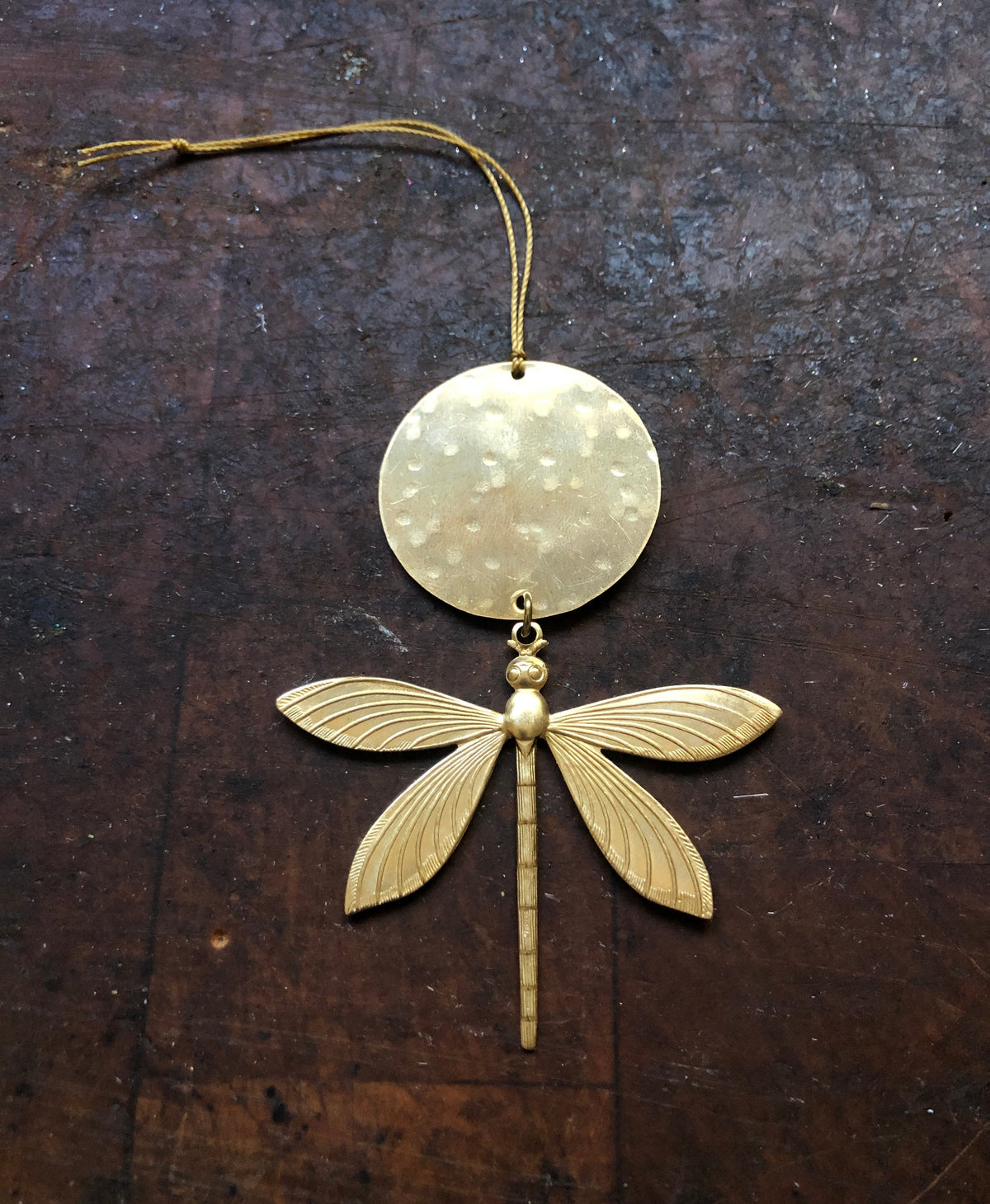 Golden metal dragonfly ornament with detailed wings suspended below a hammered circular disc, hanging by a thin gold thread on a dark textured surface.