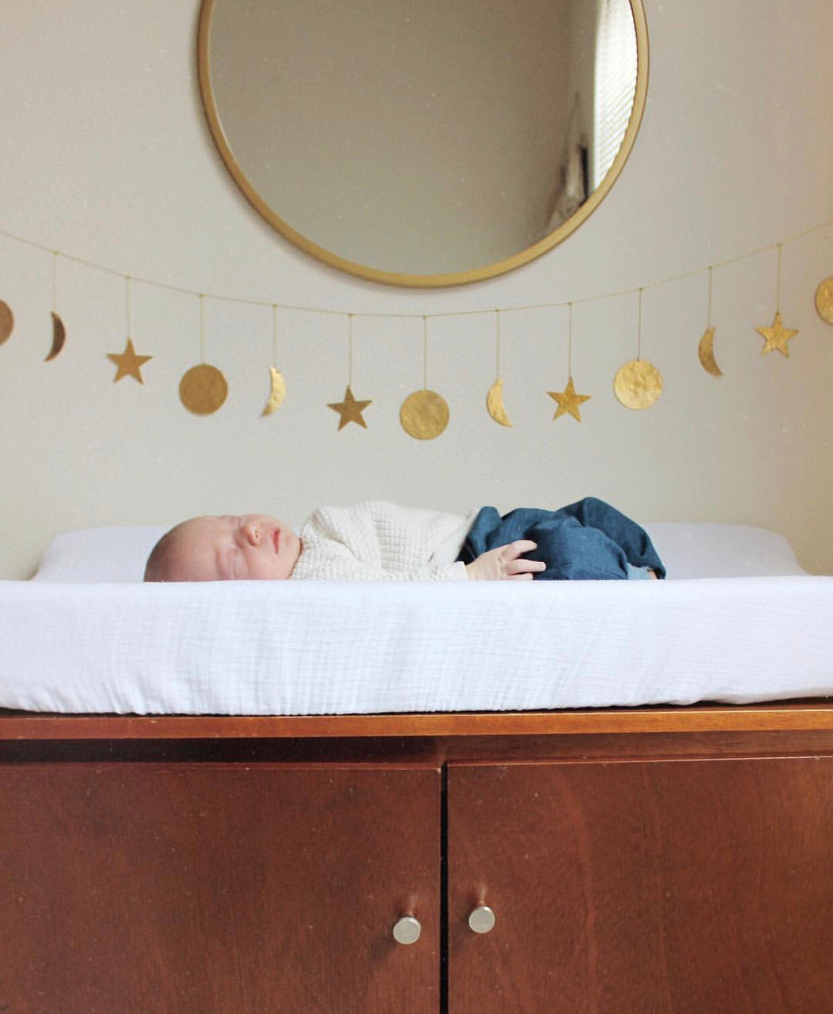 Baby lying on a white mattress with a wooden cabinet below and a circular mirror above, with a hanging brass garland featuring moons and stars on a beige wall.
