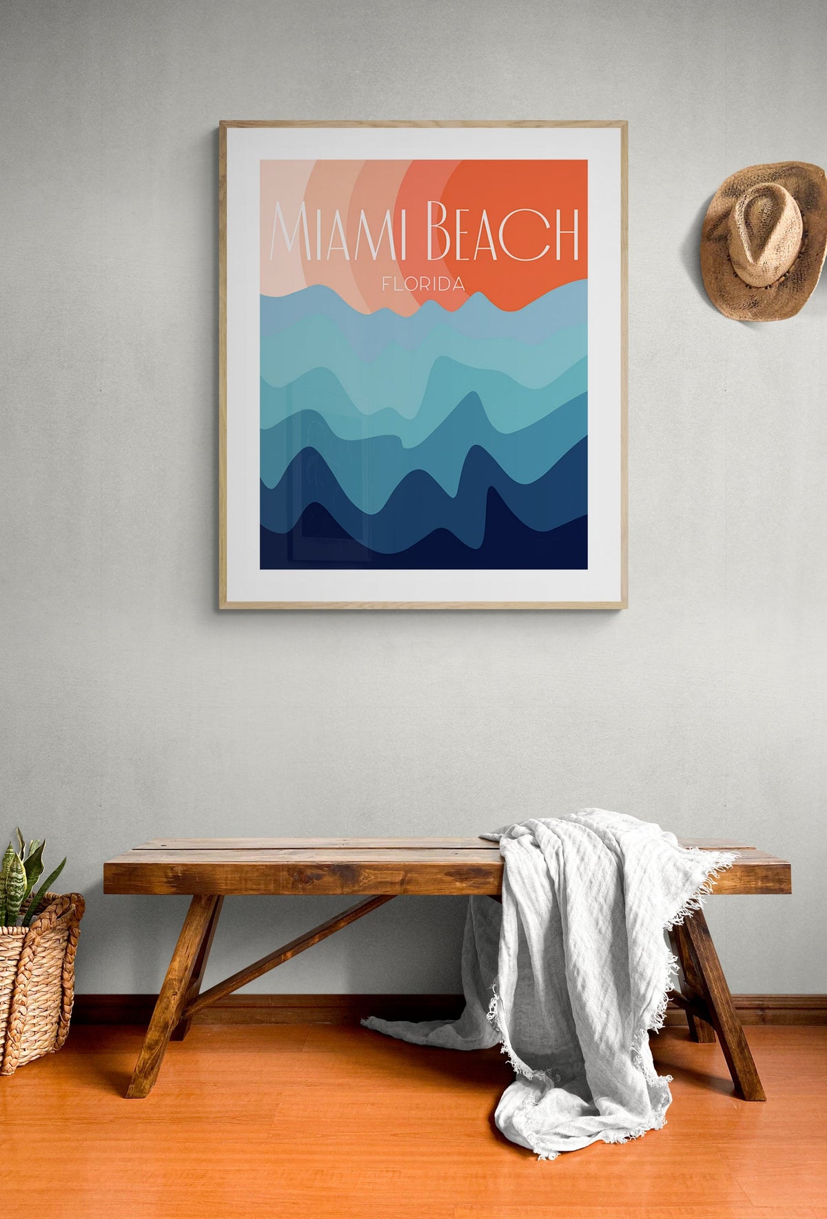 Framed modern art print with layered waves in orange, peach, and blue hues titled 'Miami Beach Florida' hanging on a light gray wall above a wooden bench with a white blanket.