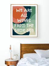 Modern art print above bed with abstract oceanic shapes in green, blue, orange hues and white text reading 'We are all waves from the same sea' in a wooden frame.