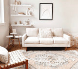 Neutral beige living room with a light beige three-seat sofa featuring a textured throw pillow, wooden legs, and a patterned area rug incorporating soft earthy tones.