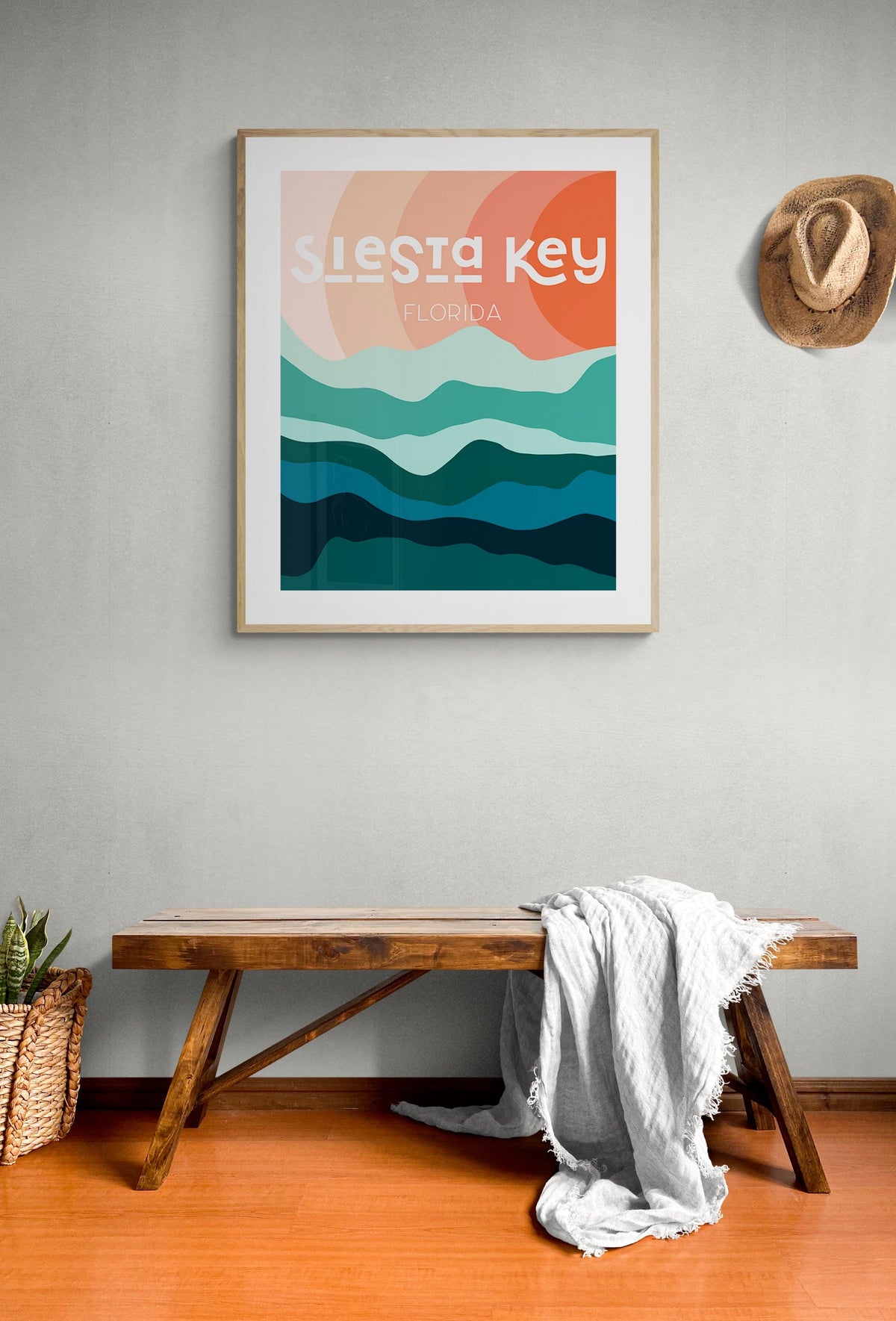 Framed modern art print with stylized wave shapes in blue and green hues under an orange sun, text reading 'Siesta Key Florida,' hanging above a wooden bench with a blanket.