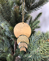 Gold-toned hammered metal ornament with four crescent shapes hanging vertically on a string, displayed on green pine branches.