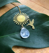 Gold sunburst pendant with a small hummingbird charm and a teardrop-shaped rose quartz stone, displayed on a large green leaf over wooden surface.