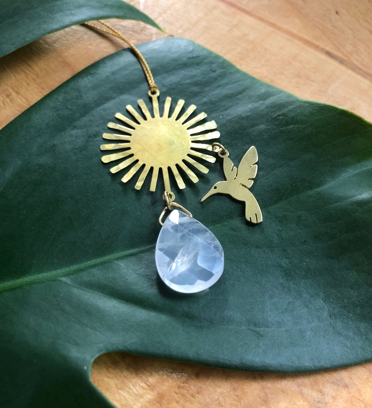 Gold sunburst pendant with a small hummingbird charm and a teardrop-shaped rose quartz stone, displayed on a large green leaf over wooden surface.
