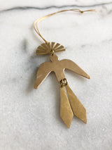 Brass bird-shaped ornament with tail feathers and a sunburst fan shape at the top, laying on a white marble surface with a hanging cord.