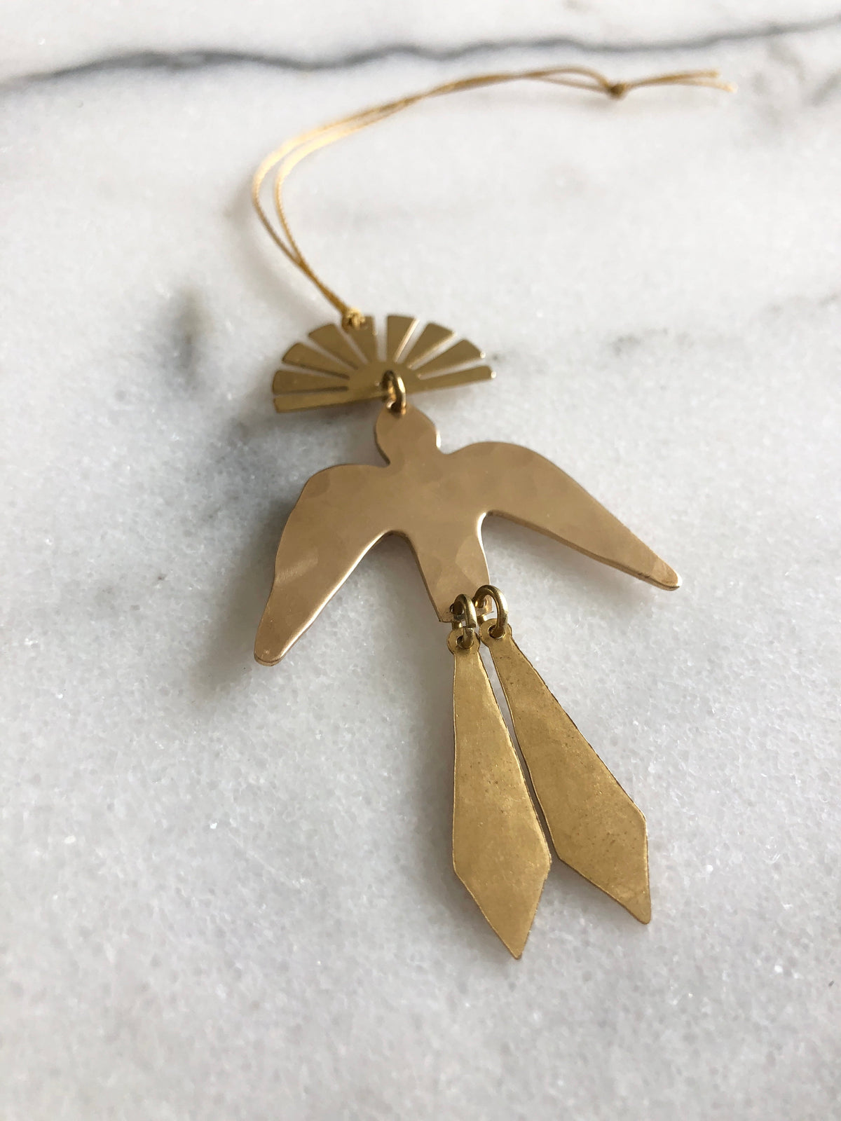 Brass bird-shaped ornament with tail feathers and a sunburst fan shape at the top, laying on a white marble surface with a hanging cord.