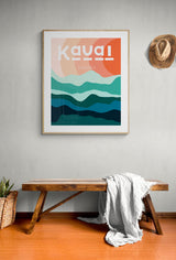 Framed modern art print with abstract hills in shades of teal and ocean blue under warm peach-orange sky, labeled 'Kauai Hawaii,' hanging on light gray wall above wooden bench.