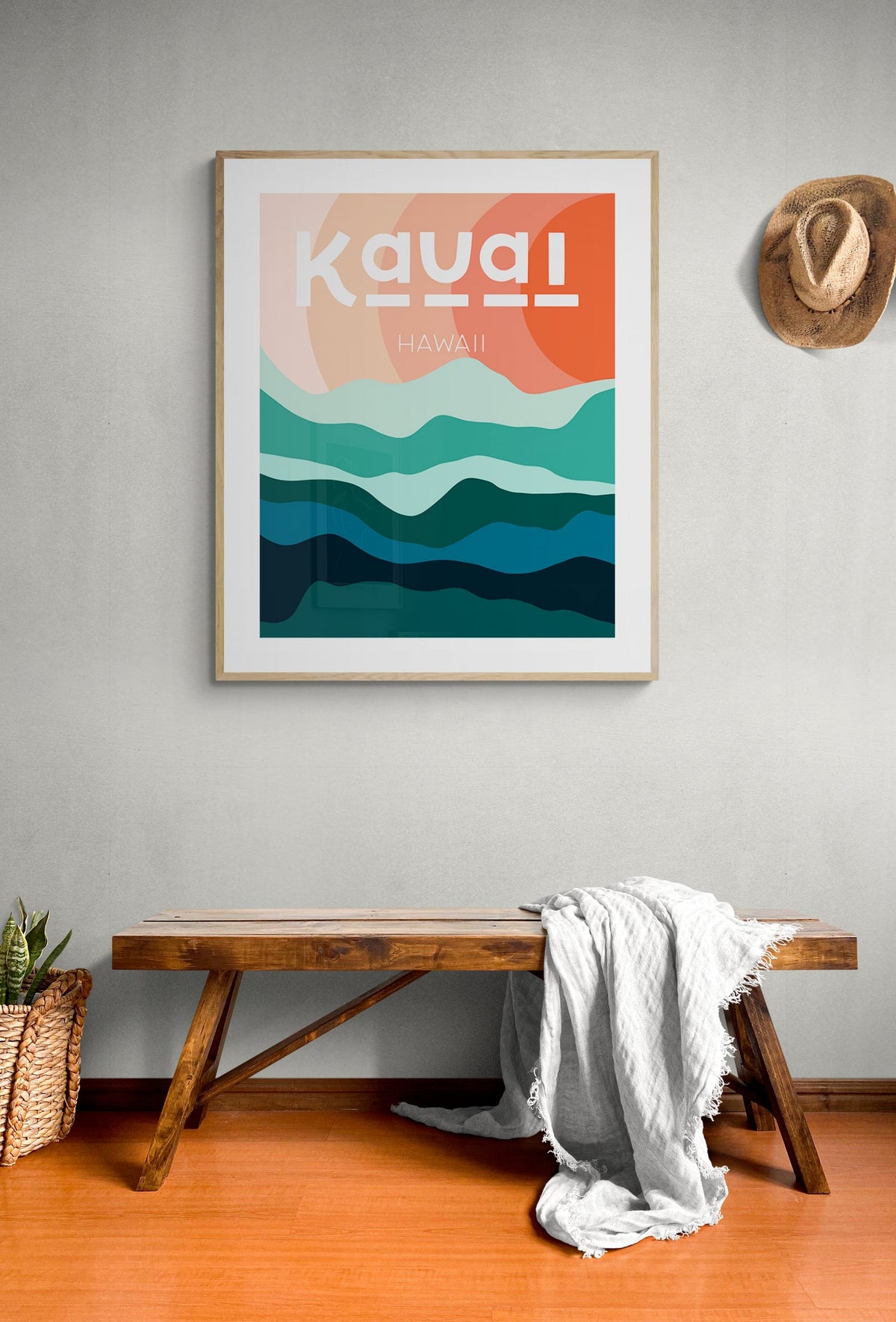 Framed modern art print with abstract hills in shades of teal and ocean blue under warm peach-orange sky, labeled 'Kauai Hawaii,' hanging on light gray wall above wooden bench.