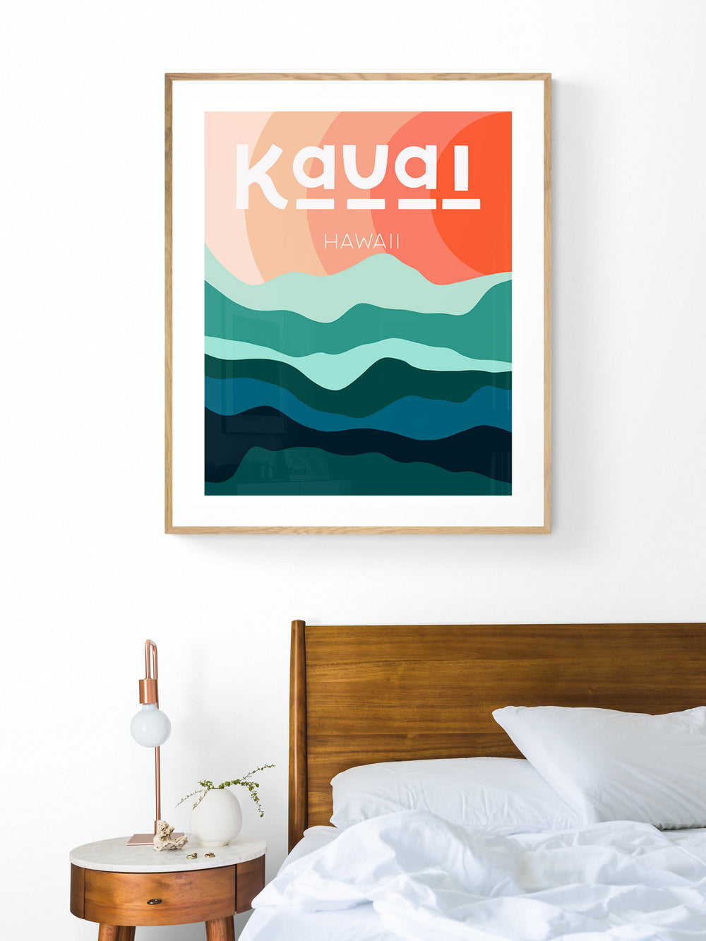 Modern art print of Kauai with layered teal and blue-green hills beneath abstract peach and orange sky, framed in light wood, hanging above wooden bed and round bedside table.