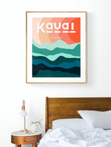 Modern art print of Kauai with layered teal and blue-green hills beneath abstract peach and orange sky, framed in light wood, hanging above wooden bed and round bedside table.