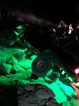 Off-road vehicle tilted on rocky terrain at night with bright green LED underglow lights illuminating rocks beneath it.