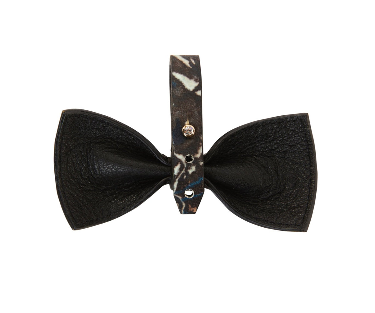 Black leather bowtie with a textured surface and a central strap featuring an abstract black and white pattern secured by two silver rivets.