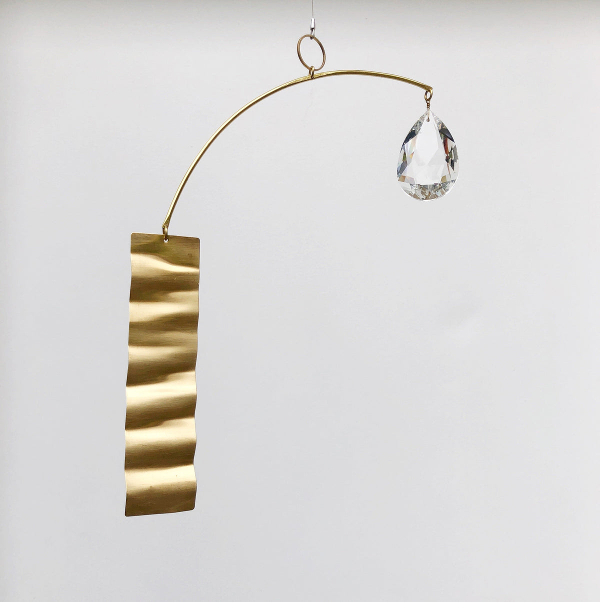 Metal suncatcher with a wavy gold rectangular strip on one end and a clear faceted teardrop crystal suspended on the other, hanging against a white background.