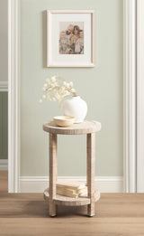 Round wooden side table with textured legs and lower shelf holding two stacked books, topped with a white vase containing light, dried flowers and a small beige bowl.