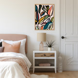 Abstract foliage artwork with bold, multicolored leaves in black, green, yellow, pink, and blue hues, framed and hanging above a white nightstand in a bedroom.