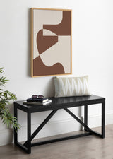 Abstract wall art with curved and angular brown and beige shapes in a natural wood frame above a black bench holding a patterned pillow, books, and sunglasses.