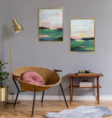 Two framed abstract coastal paintings with pastel pink, green, and blue hues hanging on a gray wall above a mid-century wooden side table and tan leather chair with a round pink cushion.
