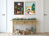 Two framed festive artworks above a wooden console table decorated with evergreen garland and candles, with firewood and a small potted pine below.