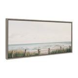 Framed canvas print depicting a calm seascape with soft blue-green water, sandy beach, grassy dunes, and a weathered wooden fence with rope, under a pale cloudy sky.