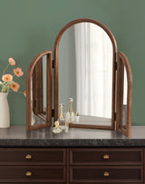 Tri-fold mirror with curved wooden frames on dark marble countertop, flanked by small glass perfume bottles and a vase of peach flowers against a sage green wall.