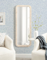 Tall rectangular wall mirror with rounded corners and a textured light wood frame, mounted on a pale blue floral patterned wallpaper between two white upholstered chairs.