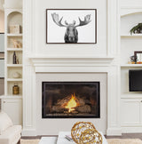 Black and white framed canvas of a moose head with large antlers centered above a white fireplace with a lit fire, flanked by built-in white shelves and cabinetry.