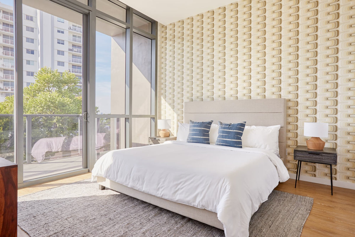 Bright bedroom with beige upholstered bed, white bedding, two indigo pillows, textured beige wall, large floor-to-ceiling windows, and wooden nightstands with white lamps.