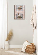 Framed canvas print of two giraffes peeking through flower-filled window boxes with pink walls, hanging on a plain light gray wall in a minimalist room.