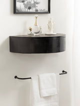 Curved black floating side table mounted on wall, decorated with glass perfume bottles and white sculptural jewelry holder, above a black towel bar with folded white towels.