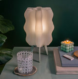 White wavy-shaped lamp with tripod legs glowing softly on a green surface, beside a patterned glass and a lit bubble candle on stacked books against a dark green background.