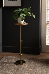 Adjustable wood and brass plant stand with a wooden circular top holding a potted green leafy plant, set against a dark wall and patterned rug.