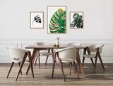 Modern dining room with a rectangular walnut table and six beige upholstered chairs, featuring three framed Monstera leaf artworks on a white wall above the table.