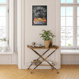 Framed chalkboard-style art with an apple pie illustration and "As American as Apple Pie" text, hanging above a wooden two-tier cart with a potted plant in a bright room.