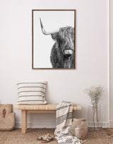 Black and white close-up art print of a Highland cow with long shaggy hair and one horn, framed and hung above a wooden bench with striped pillow and throw.