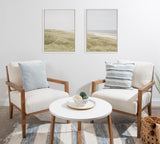 Two beige cushioned armchairs with wooden frames facing a white round coffee table, light blue and striped pillows, coastal landscape prints above, and a rug with blue and beige stripes.