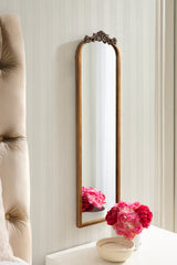 Tall rectangular mirror with an arched top and ornate carved wood detail, leaning against pale beige striped wallpaper beside a tufted beige chair and white surface holding pink flowers.