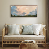 Horizontal framed canvas painting of soft pastel clouds with hints of pink, blue, and gray above green treetops, hanging above a beige cushioned bamboo-style sofa with four textured pillows.